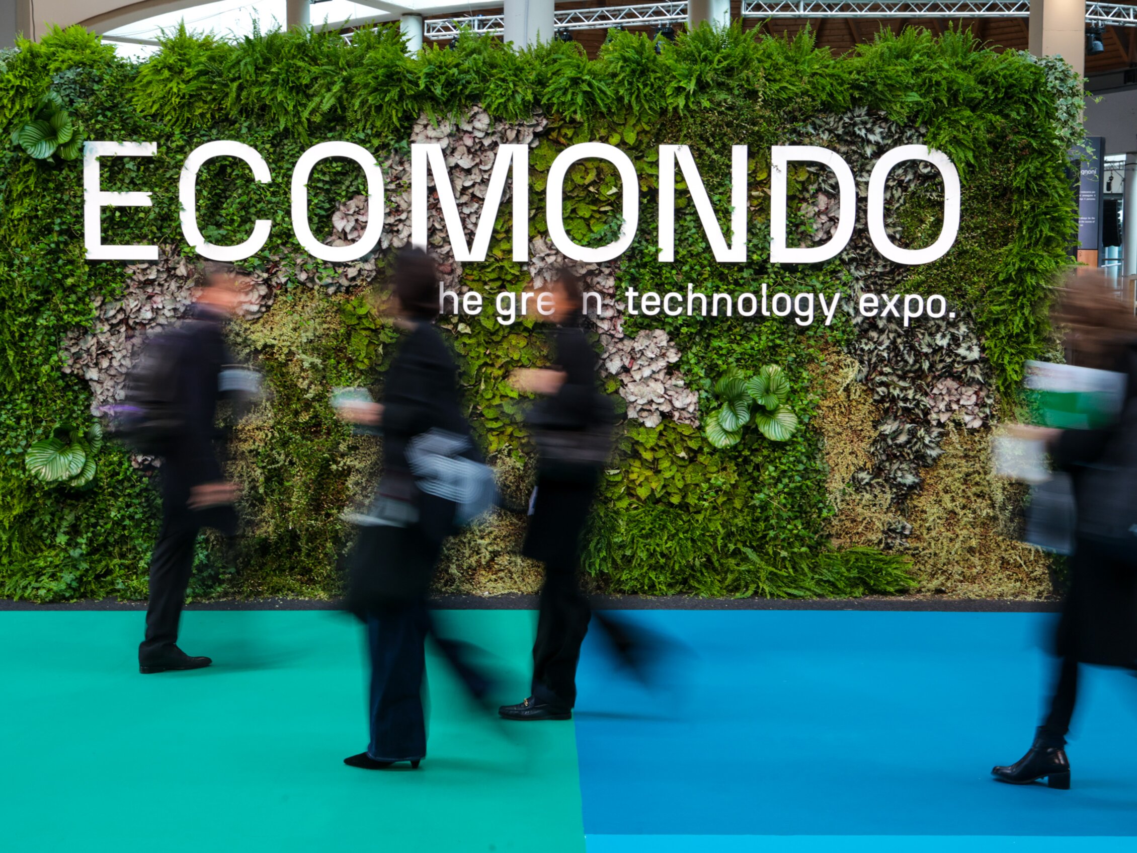 people in motion walk past a plant-covered installation with the text ECOMONDO – the green technology expo