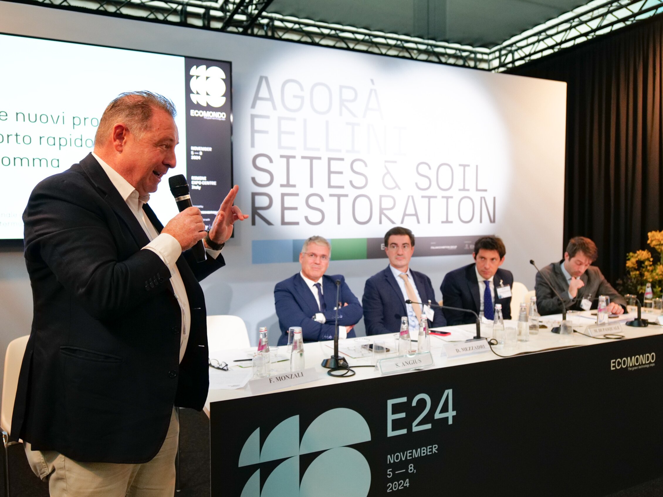 A speaker takes the floor at the 'Sites & Soil Restoration' conference, with other panelists listening