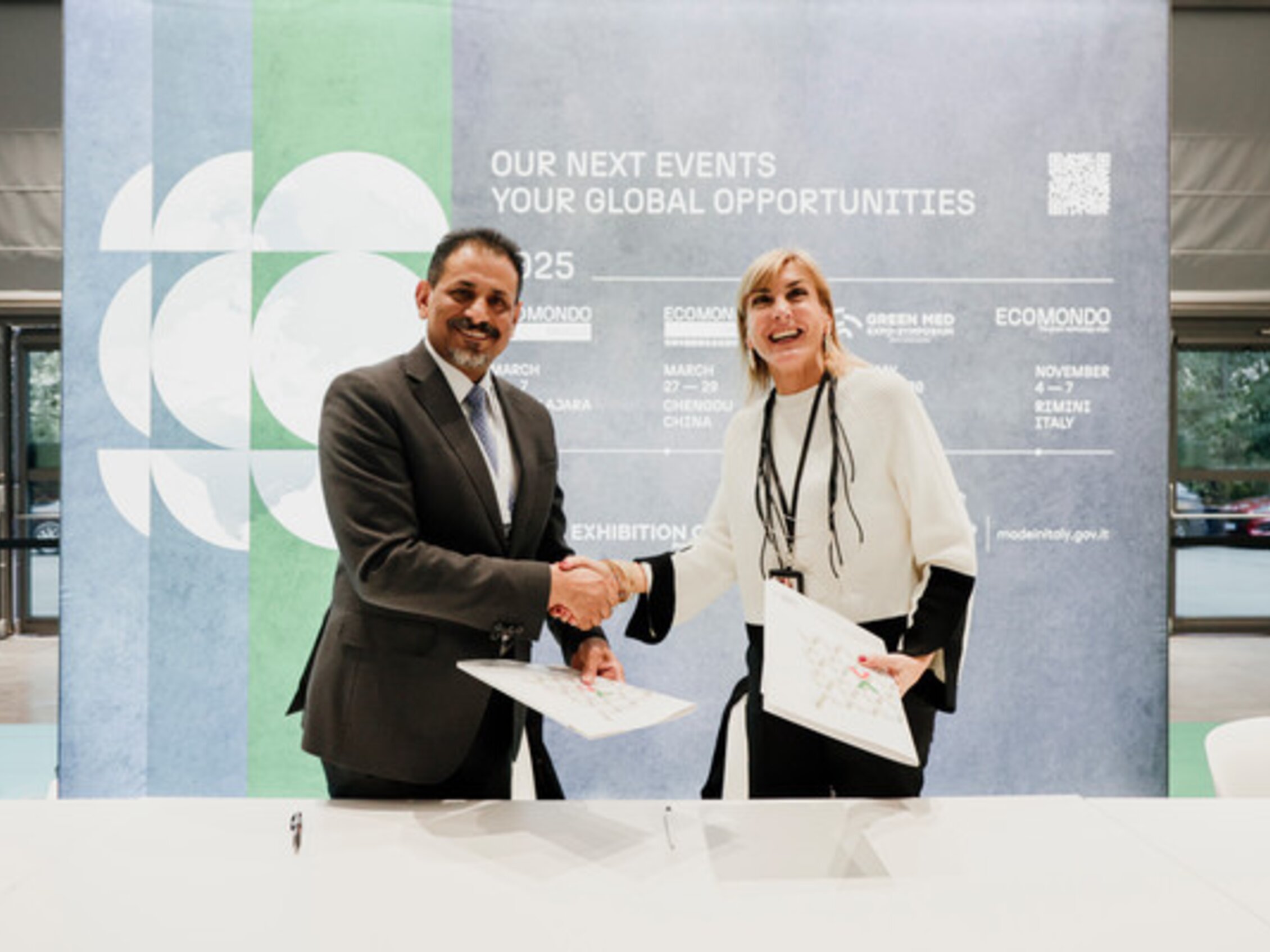 Handshake between two smiling representatives during Ecomondo, in front of a panel displaying the dates of upcoming editions.