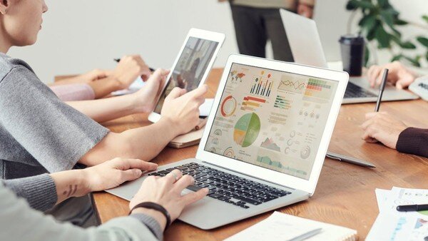 un uomo sedute ad un tavolo consulta grafici e schemi al pc mentre altre persone lavorano attorno