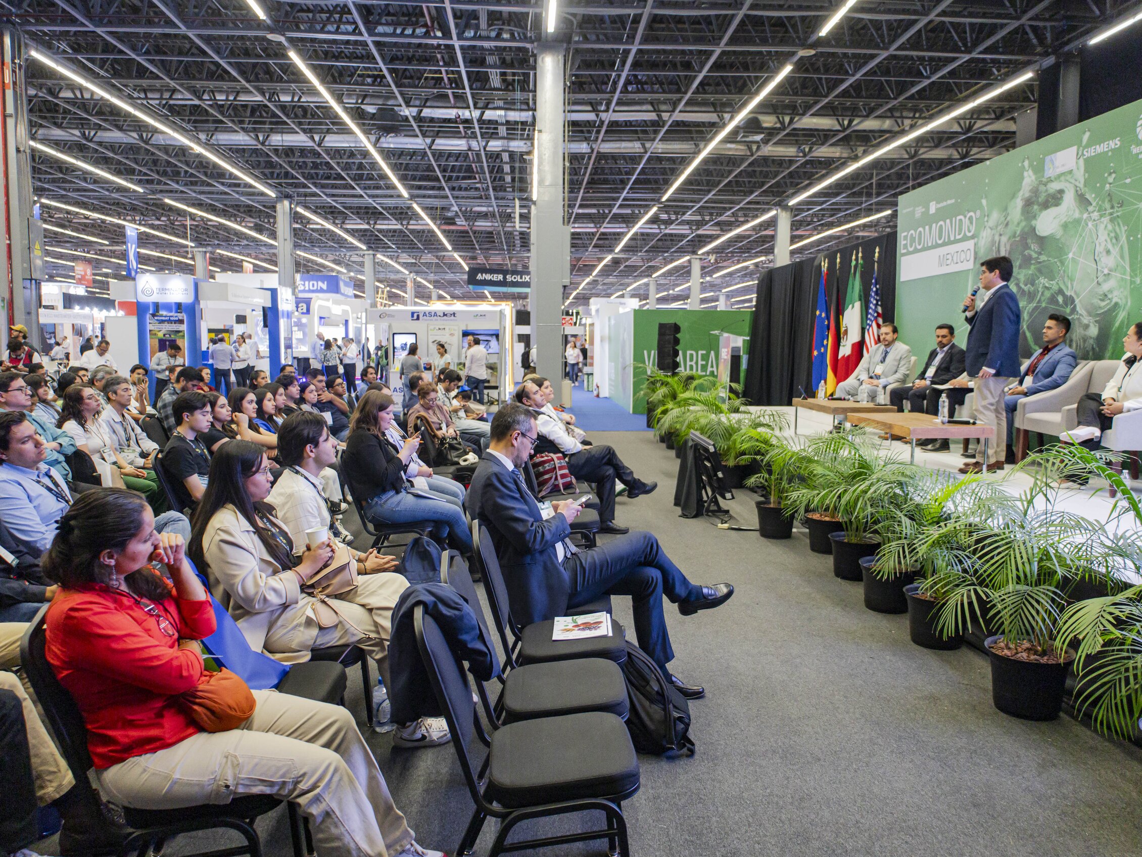 Italiano: Una conferenza all'interno di un ampio spazio espositivo, con un relatore sul palco che parla a un pubblico seduto. Sullo sfondo, un grande schermo verde con la scritta 'ECOMONDO' e stand espositivi