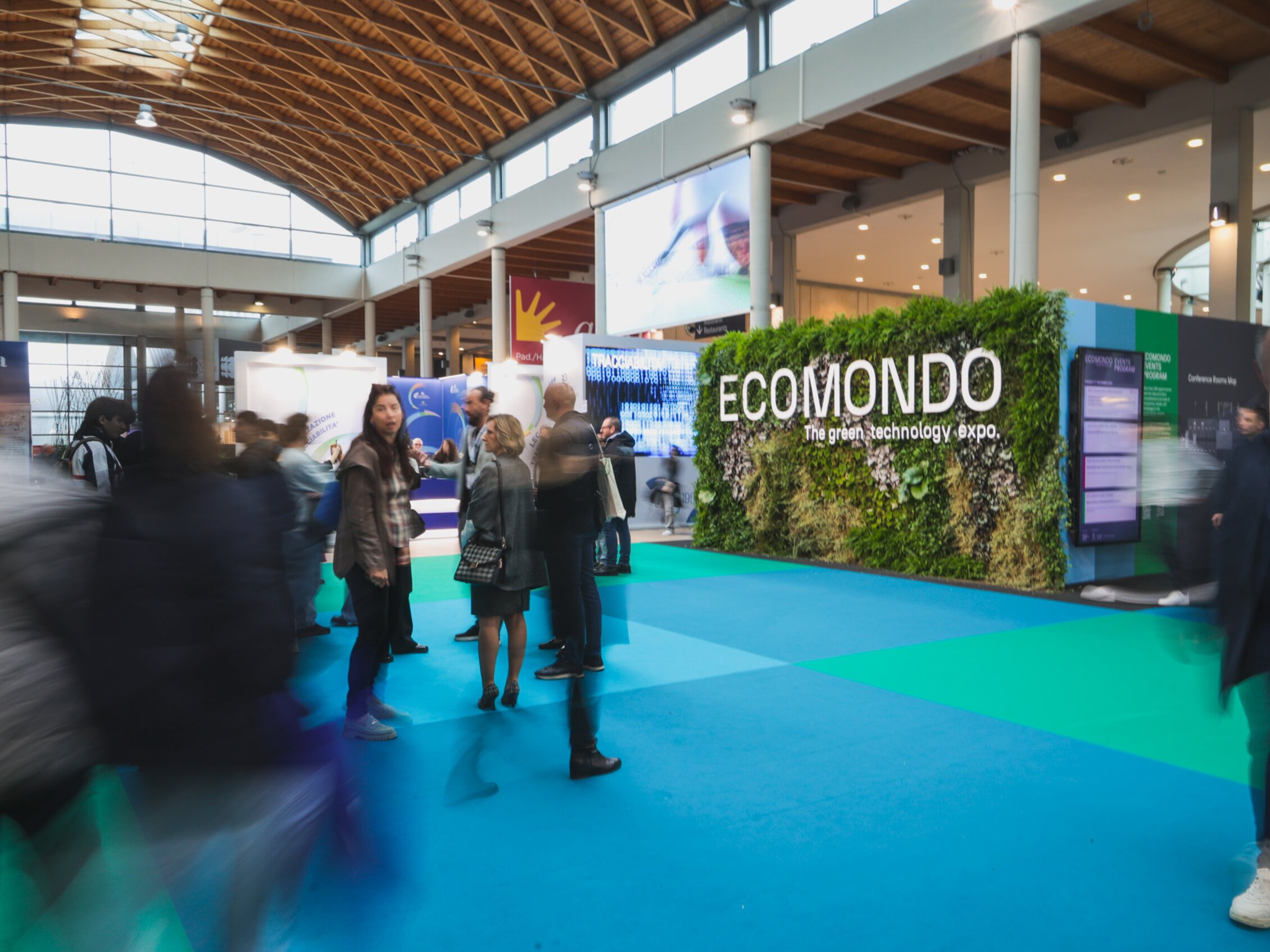 Numerose persone camminano tra gli stand della fiera, dove spicca una grande installazione verde ricoperta di piante con la scritta 'ECOMONDO - The green technology expo' al centro